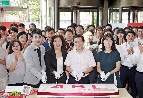ABL생명 사명변경 1주년을 맞아 순레이 사장과 입직원 들이 기념 케이크를 자르며 밝게 웃고 있는 모습, 그 주위로 수많은 직원들이 박수를 치고 있는 사진